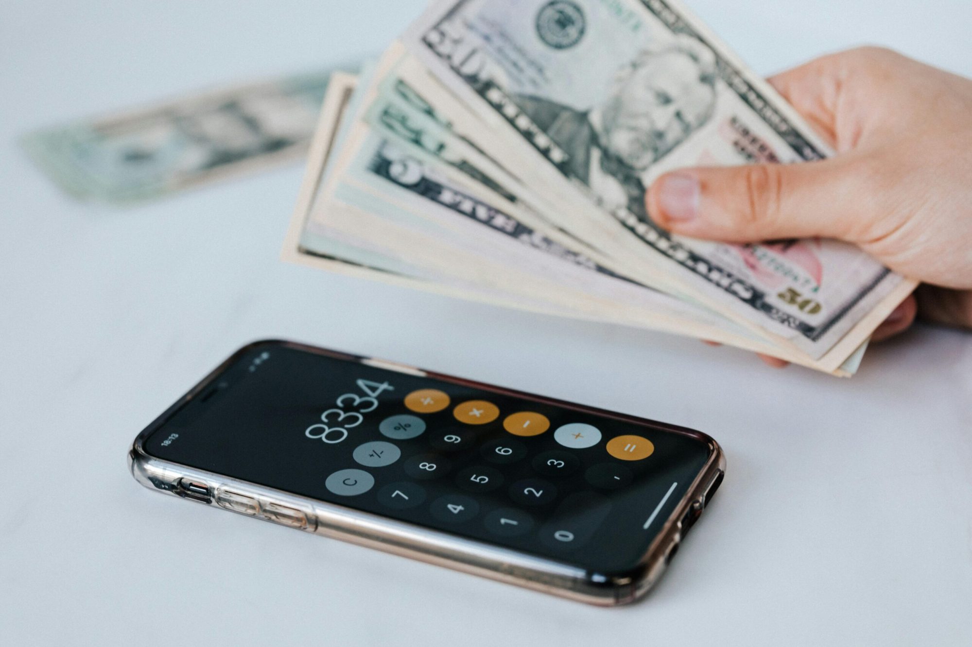pexels-photo-4386292-4386292 Calculator application on mobile phone screen on white surface near crop anonymous person holding pile of dollars